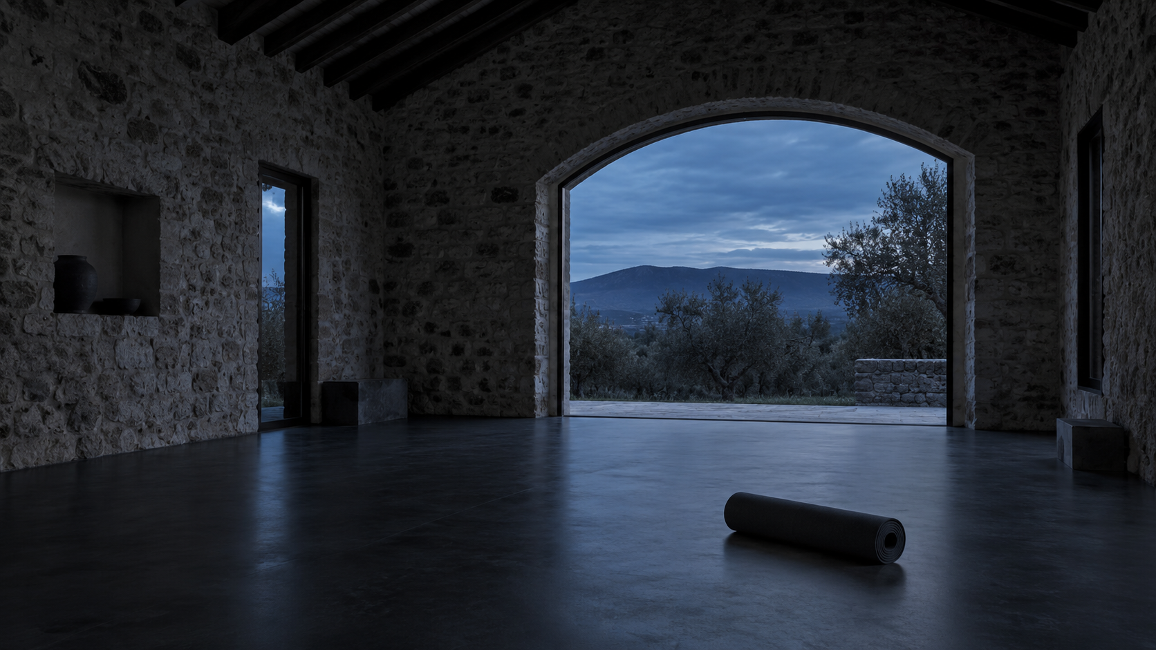 Interior de una antigua cuadra rehabilitada como sala de yoga, con los muros de piedra originales a la vista y luz azulada entrando por el vano abierto al olivar