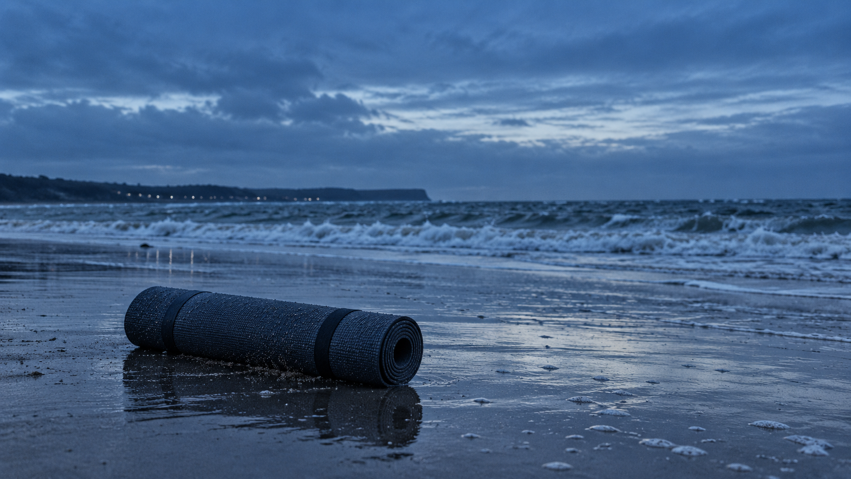 Esterilla de yoga enrollada sobre la arena de una playa atlántica al amanecer, con el mar picado al fondo y nadie alrededor