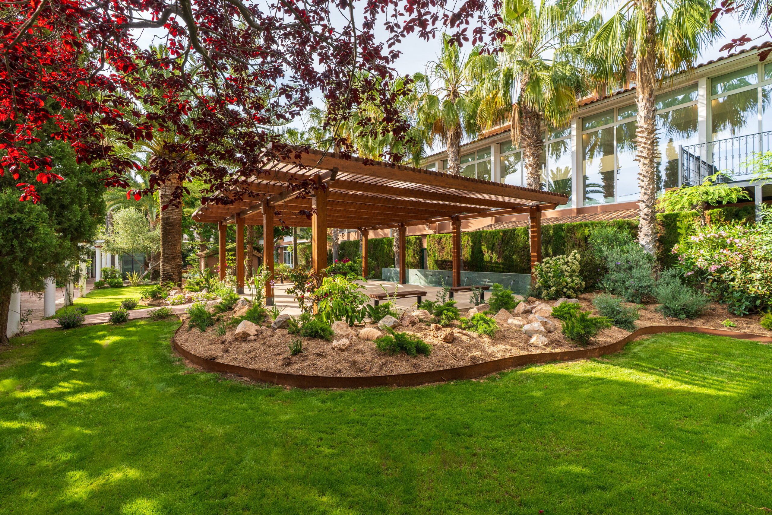Jardín de Palasiet con una pérgola de madera entre palmeras, césped y la galería acristalada de la villa al fondo