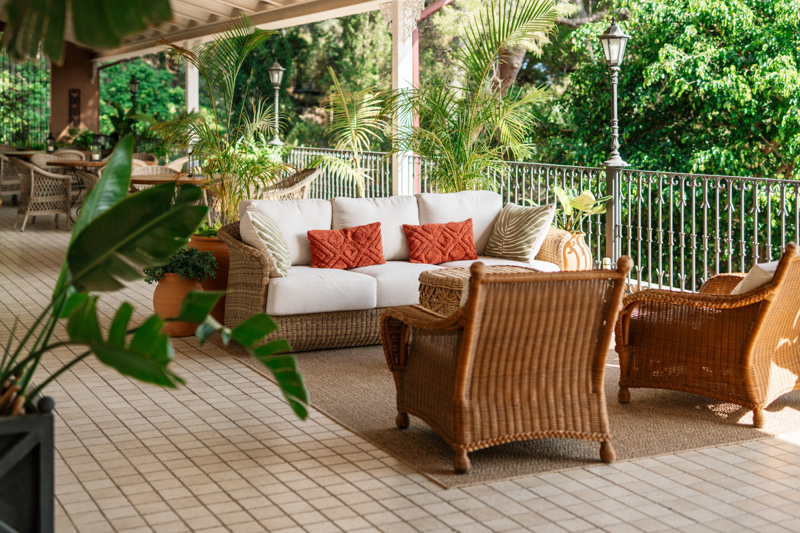 Terraza cubierta de Palasiet con un sofá y butacas de mimbre, plantas y una barandilla de forja ante los árboles